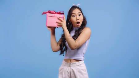 schockierte junge asiatische Frau schüttelt umhülltes Geschenk isoliert auf blau
