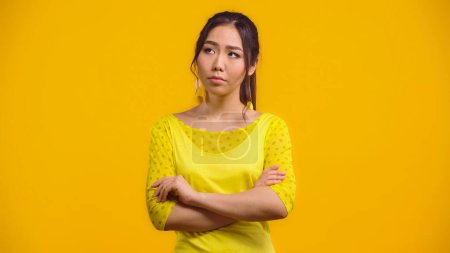 worried and young asian woman standing with crossed arms isolated on yellow 