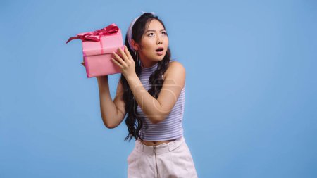puzzled asian woman shaking wrapped gift box isolated on blue