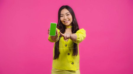 feliz asiático mujer apuntando a smartphone con verde pantalla aislada en rosa  