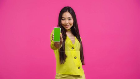 happy asian woman holding smartphone with green screen isolated on pink 