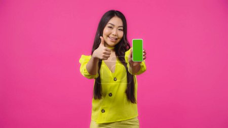 happy asian woman holding smartphone with green screen while showing thumb up isolated on pink 