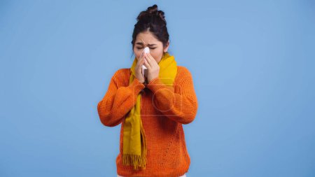 kranke Asiatin in orangefarbenem Pullover und Schal niesen in Serviette isoliert auf blau 