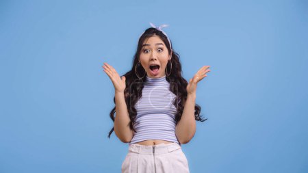 amazed and young asian woman with opened mouth gesturing isolated on blue 