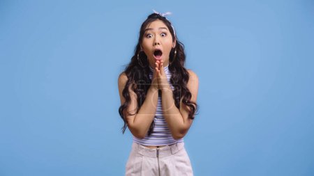 shocked and young asian woman with opened mouth and praying hands isolated on blue  