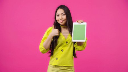 fröhliche asiatische Frau hält digitale Tablette mit grünem Bildschirm und zeigt sich wie isoliert auf rosa 