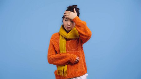 mujer asiática enferma en suéter naranja y bufanda tocando la frente aislada en azul 