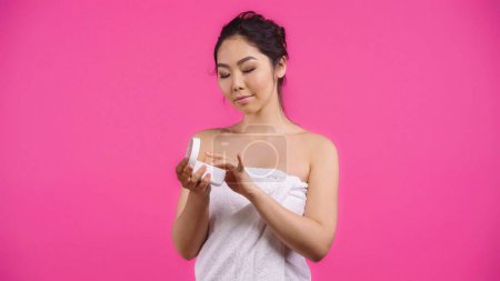 Téléchargez les photos : Asiatique jeune femme dans serviette tenant conteneur avec visage crème isolé sur rose - en image libre de droit