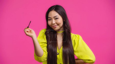cheerful asian woman holding mascara applicator and smiling isolated on pink 