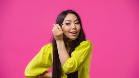 Téléchargez les photos : Gai asiatique femme appliquer mascara et sourire isolé sur rose - en image libre de droit