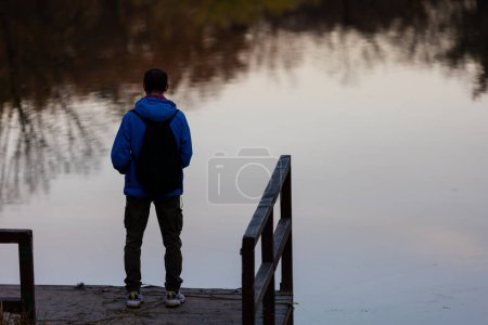 Photo for Young man with backpack standing alone on wooden footbridge and staring at lake. Peaceful atmosphere in nature. Enjoying fresh air in autumn evening. Back view. - Royalty Free Image