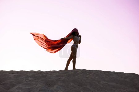 Silhouette einer Frau, die lange rote Stoffe auf Sand unter rosa Himmel hochhält, Inspiration