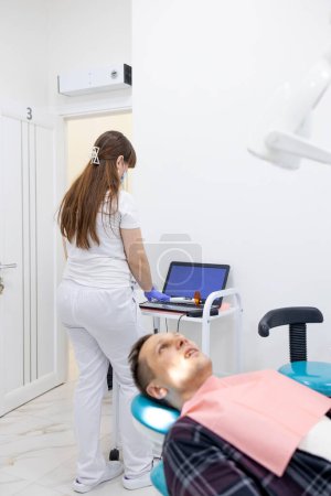 Téléchargez les photos : Dentiste utilisant un ordinateur portable pendant qu'un collègue examine un patient masculin à la clinique dentaire. - en image libre de droit