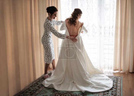 Téléchargez les photos : Une magnifique mariée blonde dans une robe de luxe blanche se prépare pour le mariage. Préparation du matin. Une femme met une robe. - en image libre de droit