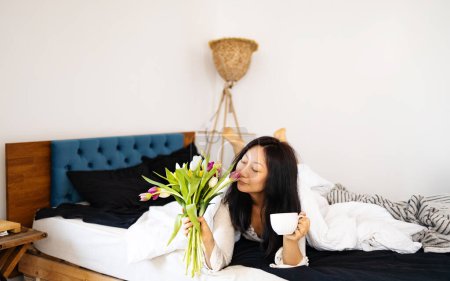 Young Asian woman holding cup and tulips flowers in the bed. Wake up morning concept.