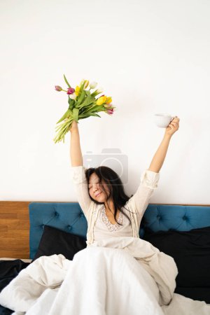Young Asian woman holding cup and tulips flowers in the bed. Wake up morning concept.