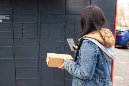 Photo for Young woman using smart phone while standing with a parcel delivered with post office machine with automatic lockers. New technologies in delivery service, self picking - Royalty Free Image
