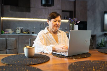 Ältere Frau im Hemd blickt auf Laptop-Bildschirm und lächelt in moderne Küche.