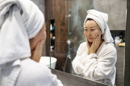 Portrait of beautiful Asian girl in bathrobe and with towel on her head cleaning her face and smiling in bathroom.