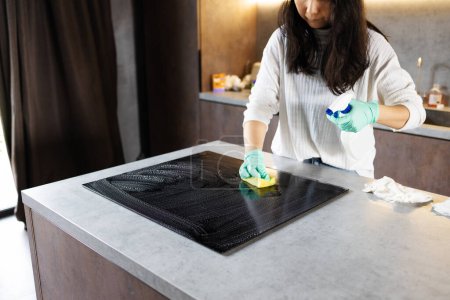 Close-up Of Person Hands Cleaning Induction Stove In Kitchen With Spray Bottle And Sponge.
