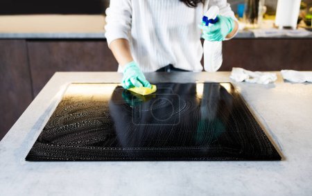 Close-up Of Person Hands Cleaning Induction Stove In Kitchen With Spray Bottle And Sponge.