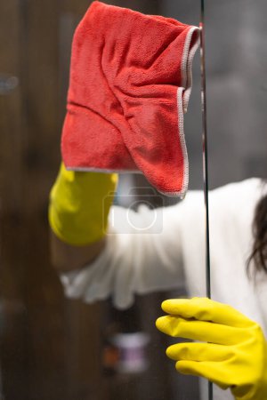Téléchargez les photos : Gros plan d'une femme en gants jaunes nettoyant une porte en verre dans la salle de bain. - en image libre de droit