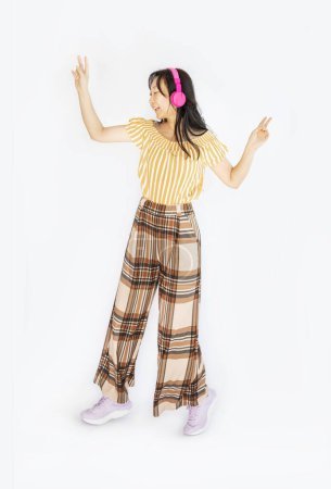 Full size body length studio portrait jump shot of glad cheerful joyful lovely Asian girl with headphones jumping up wearing trendy outfit isolated on white background.