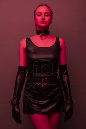 Foto de Beautiful young woman in a leather dress and bondage set posing on red light backgound - Imagen libre de derechos