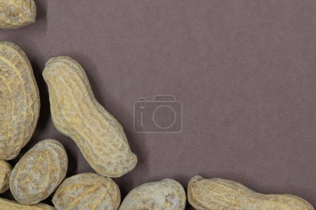 Unshelled peanuts scattered on brown cardstock background with copy space. Flat lay closeup food, legume crop nutrition and allergy, farming industry concept.