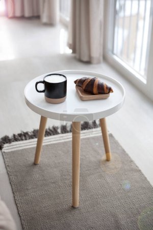 Foto de Mesa con croissant, taza y sillón suave en sala de estar ligera - Imagen libre de derechos