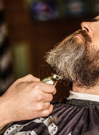 Barbudo en la barbería. Peluquero trabaja cortador de barba. Hipster cliente cortarse el pelo. Manos de peluquero con un cortador de barba, primer plano.