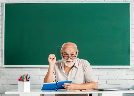 Photo for Portrait smiling man in glasses sit at desk. Old man teachers on green board. Happy school male teacher book. Portrait male teacher in classroom. Man elementary school teacher standing in classroom. - Royalty Free Image