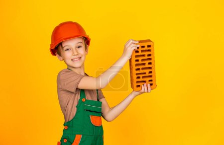 Photo for Little builder in helmet. Child dressed as a workman builder. Boy wearing helmet. Child building helmet, hard hat. Boy in a construction helmet holds a brick in his hands on yellow background. - Royalty Free Image