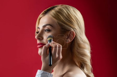 Make up in process. Girl hold blush blusher apply powder visage isolated red background. Young woman powdering cheeks. Makeup brush. Hand of visagiste, painting cosmetics beauty model girl.