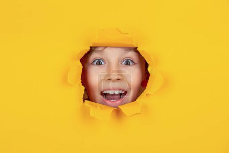 Kid with toothy smile shows face in paper hole. Portrait of beautiful happy smiling little boy posing in yellow paper hole. Banner for discounts and sales. Positive child toothy pleasant smile on face