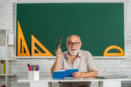 Photo for Smiling man in glasses sit at desk. Old man teachers on green board. Happy school male teacher book. Portrait male teacher in classroom. Man elementary school teacher standing in classroom. - Royalty Free Image