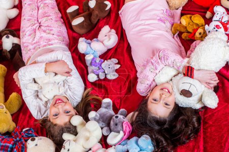 Photo for Girl sisters in pajamas lie in bed, top view. Children in soft warm pajamas playing home. Girls in colorful pajamas hold funny stuffed animals, childhood, happiness. - Royalty Free Image