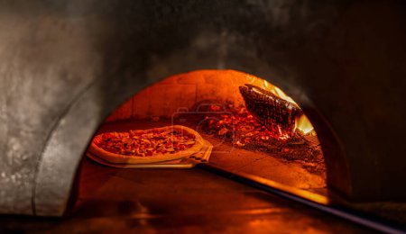 Pizza-Konzept. Zubereitung traditioneller italienischer Pizza. Schaufel für Pizza, Teig backen im Ofen mit offenem Feuer. Pizzaiolo bei der Überprüfung einer im Holzofen gekochten Pizza mit seiner Ofenschaufel.