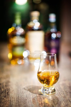 Foto de Color de cerca disparó un vaso de whisky Glencairn en una mesa de madera, con poca profundidad de campo, y botellas en el fondo. - Imagen libre de derechos