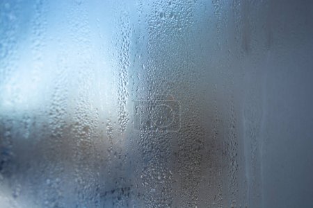 A foggy window, a glass surface covered with small drops of water. Through the glass you can see a blurred background with shades of blue and gray. The image conveys an atmosphere of coolness and moisture