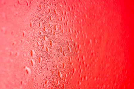 The photo shows a close-up of a surface covered with water droplets with a red background. The drops are arranged randomly and have different sizes, creating an interesting texture and rich picture. The warm red background gives the image energy and 