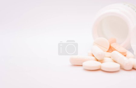 Scattered pills and capsules falling out of an open white container are shown on a white background. Medicines of different shapes and colors: round, oval and capsules, mostly white and orange shades