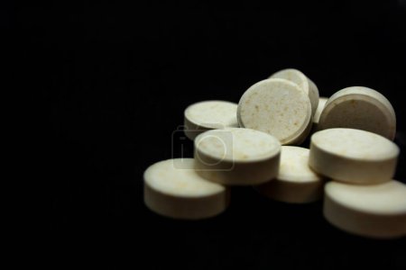 A pile of round, light-yellow tablets against a black background. The tablets have small specks and are arranged closely together.
