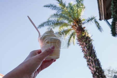 Téléchargez les photos : Hand holds up a cup of soft serve pineapple vanilla swirl ice cream, with palm trees in the background - en image libre de droit