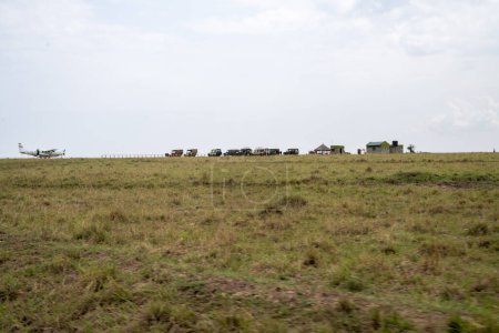 Foto de Kenia, África - 10 de marzo de 2023: Vista lejana de la pista de aterrizaje de Musiara en la reserva de Masaai Mara, con un pequeño avión arbusto y vehículos de safari esperando para recoger a los turistas - Imagen libre de derechos