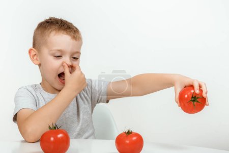 Foto de Al chico no le gusta el tomate. Concepto de nutrición y hábitos alimenticios saludables para los niños. A los niños no les gusta comer tomates rojos. Pequeño niño lindo, chico se niegan a comer verduras saludables. - Imagen libre de derechos