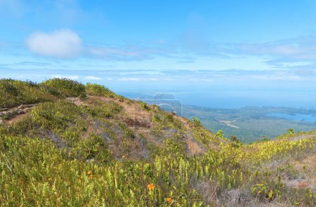 Grüne Hügel und orangefarbene Blumen überragen ein riesiges Gewässer unter einem klaren Himmel mit verstreuten Wolken. Mombacho Volcano Natural Reserve, Nicaragua.