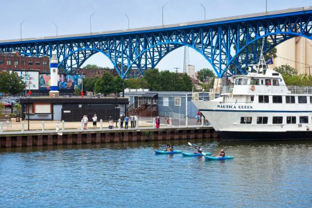 Photo for Cleveland, OH - July 3, 2022: Three kayakers paddle past the Nautica Queen, a popular excursion boat on the Cuyahoga River in downtown Cleveland, Ohio, with the Main Avenue bridge passing overhead. - Royalty Free Image