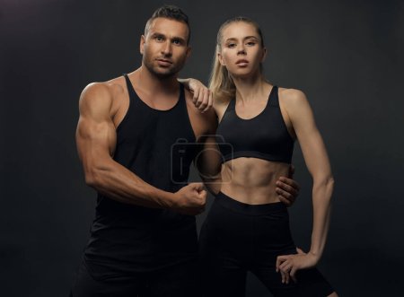 Photo for Muscular man and fitness girl standing together in studio. Portrait of two health people in sport outfit. Sport couple in black sportswear. Handsome hunk and beautiful woman with six pack abs - Royalty Free Image