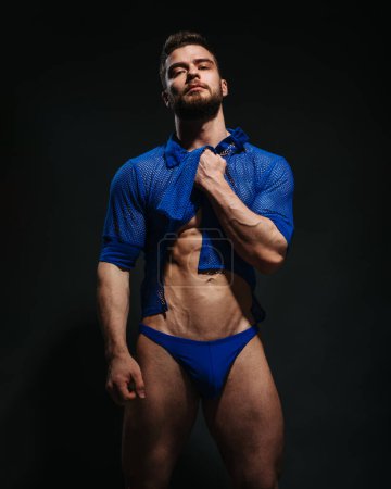 Photo for Sexy muscled male model in blue shirt and blue swimwear on black background. Bearded man taking his clothes up. Fashion guy in beachwear in studio. Male fitness model in stylish outlook. - Royalty Free Image